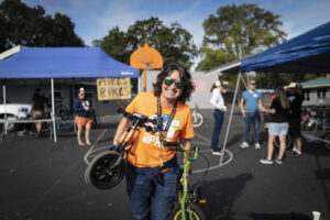 Cara is smiling as she walks with 2 kids bikes in her arms. She is at a community event and wearing an orange Bike PHE shirt.