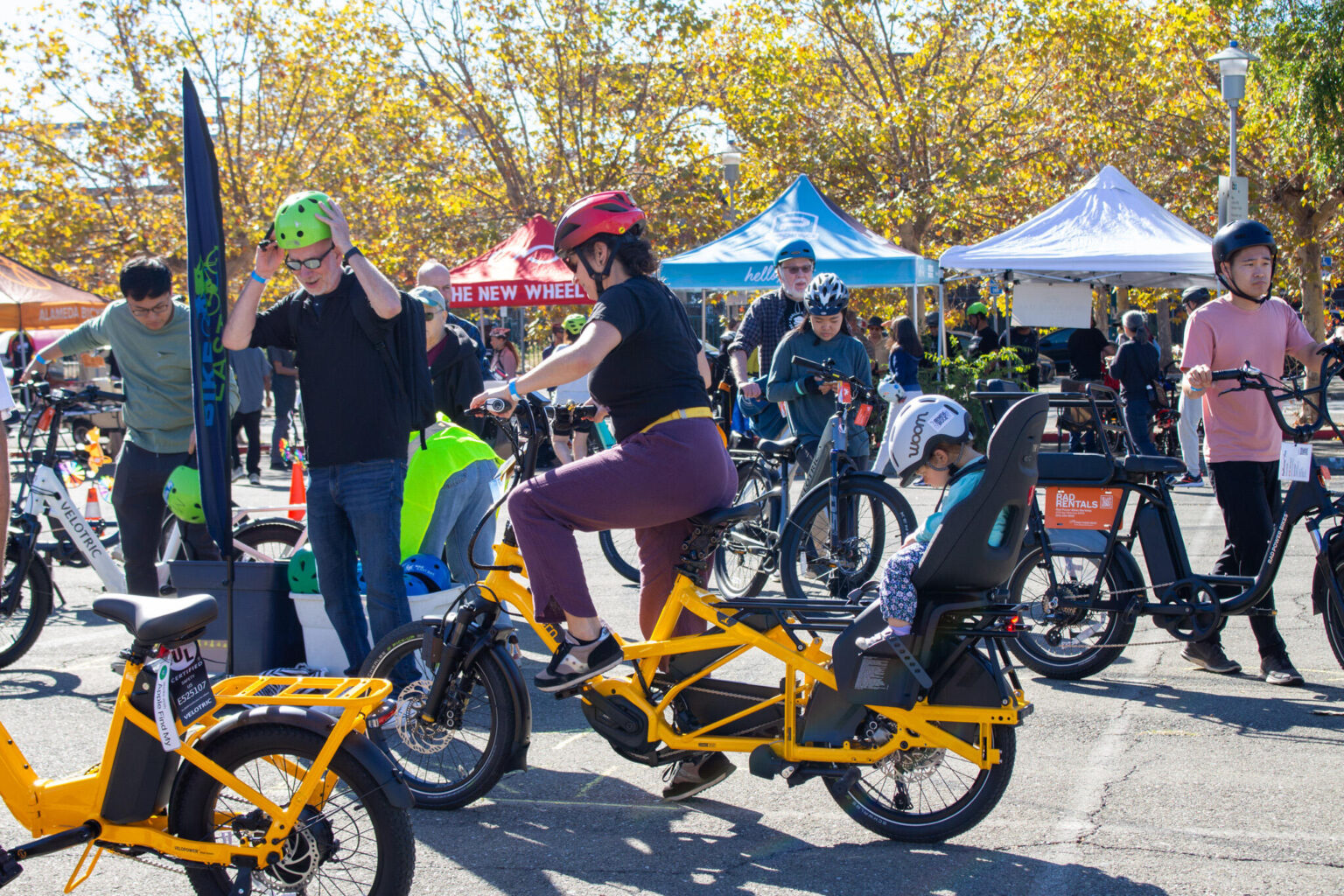 An outdoor community event showcasing e-bikes and cargo bikes. A large crowd is gathered among display tents. In the foreground, a woman wearing a red helmet sits on a bright yellow cargo bike with a child passenger seat, while a man in a green helmet stands next to a similar yellow e-bike. Many other people are visible inspecting the bikes and walking around on the paved ground, shaded by trees.