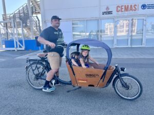 Justin is riding on a long cargo bike, with 2 kids riding inside a covered bucket seat attached. They are looking at the camera smiling, and 1 child is sleeping.