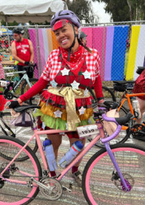 Kariz is dressed super colorfully, with a rainbow tutu skirt and a cowboy themed top and bandana. She is standing at a race with pride flags in the background, and a fully pink and purple bike.
