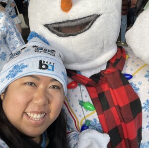 Kendra is smiling alongside a snowman mascot. Both are decked out in BART Christmas gear.
