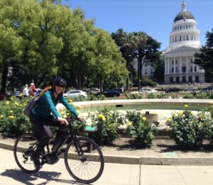 Melanie is smiling and riding her bike in front of the Capitol building and rose garden.