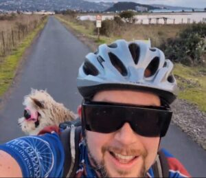 Pat is riding down a scenic nature trail, taking a selfie that includes him in his bike gear, and a small dog wearing riding goggles riding in his backpack.
