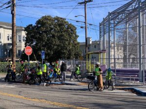 A street-level photograph showing a large group of children and adults participating in a "Bike Bus" on a sunny day in Berkeley. Participants: Numerous people are wearing bright yellow high-visibility safety vests and bicycle helmets. Many are mounted on bicycles or standing near them at a street corner. Location: The group is gathered near a stop sign and a tall chain-link fence belonging to a sports court or park area. Residential buildings and large green trees are visible in the background under a clear blue sky.