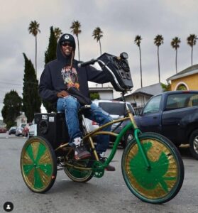 RB is riding a cruiser bike with a sound system attached and colorful taped up wheels. Palm trees are in the background.