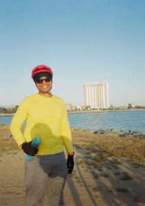 Rickie is wearing a neon yellow shirt and red helmet, smiling as he stands in front of a body of water.